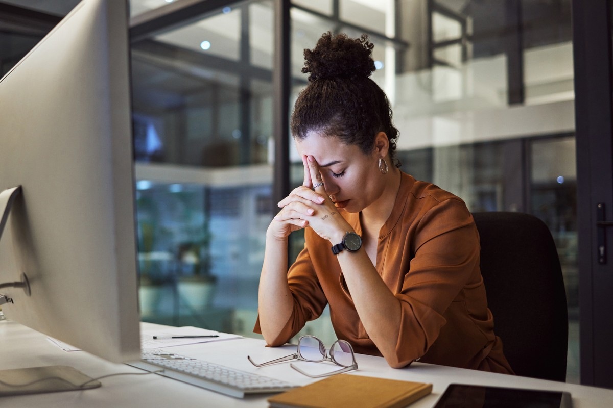 Stressed woman at a desk in an office, hands pressed to her forehead while looking at a computer screen.